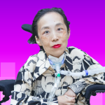 Photo of Alice Wong, an Asian American disabled woman in a power chair. She is wearing a black blouse with a floral print, a bold red lip color and a trach at her neck. Photo credit: Eddie Hernandez Photography.