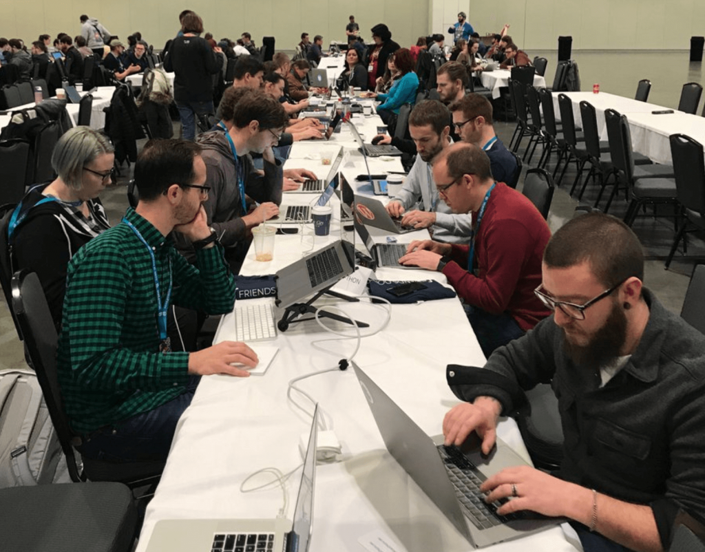 25 developers sit at a long table, each with their laptops, working on various WordPress accessibility projects. Ten developers in the foreground huddled, working with Deque’s axe.
