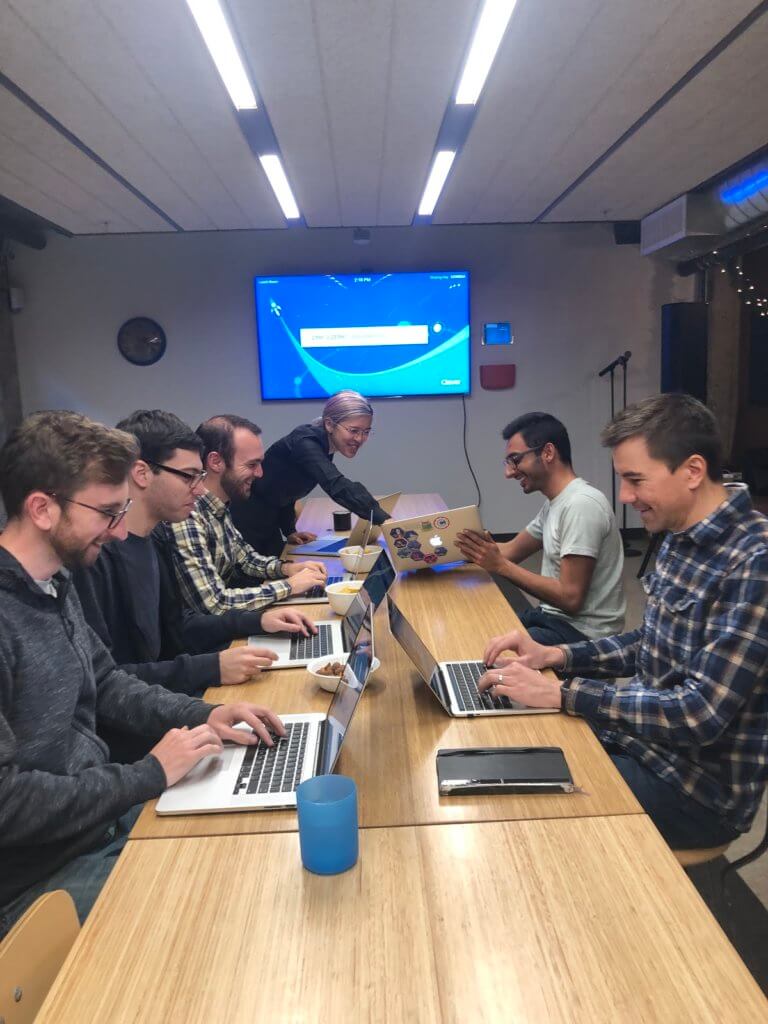 Six developers seated around a table, cheerfully working on laptops.