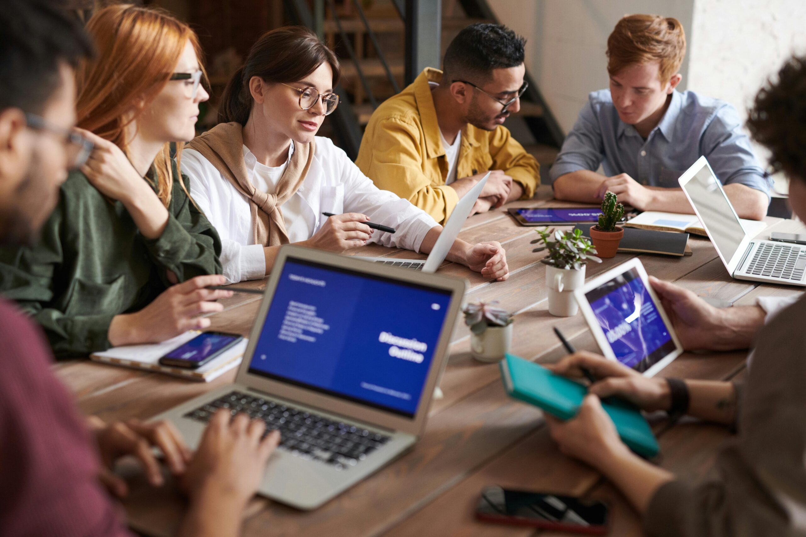 group of people sitting at a conference table with computers, tablets, and note pads