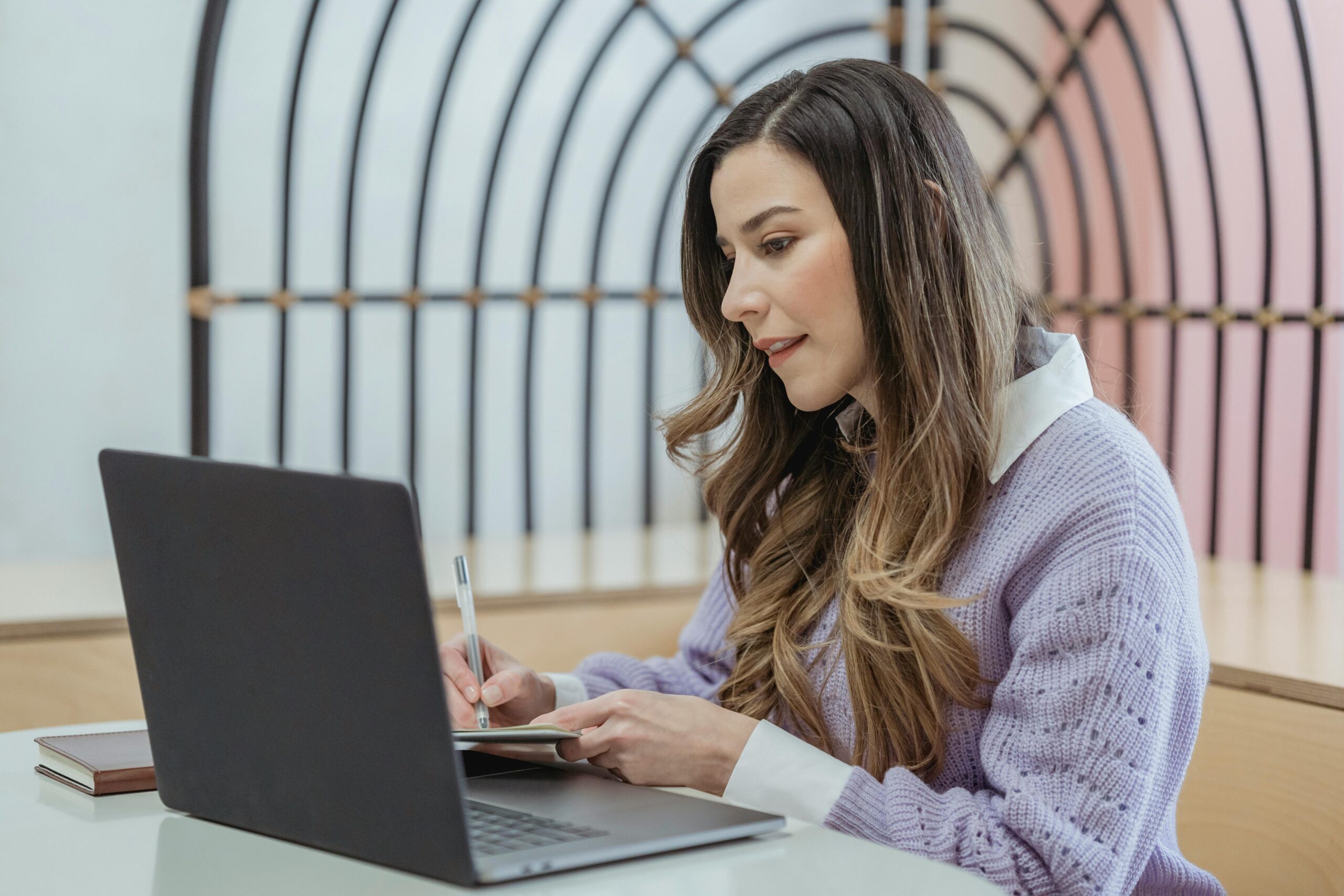 A woman looking at laptop screen and taking notes on a notepad.