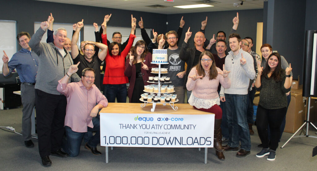A picture taken in the Deque office in Ann Arbor, Michigan, back in 2018, when a group of Deque employees met to celebrate the first one million downloads of Axe-core. The team is smiling and cheering as they stand around a desk with branded cupcakes and a sign thanking the A11y community for 1 million downloads.